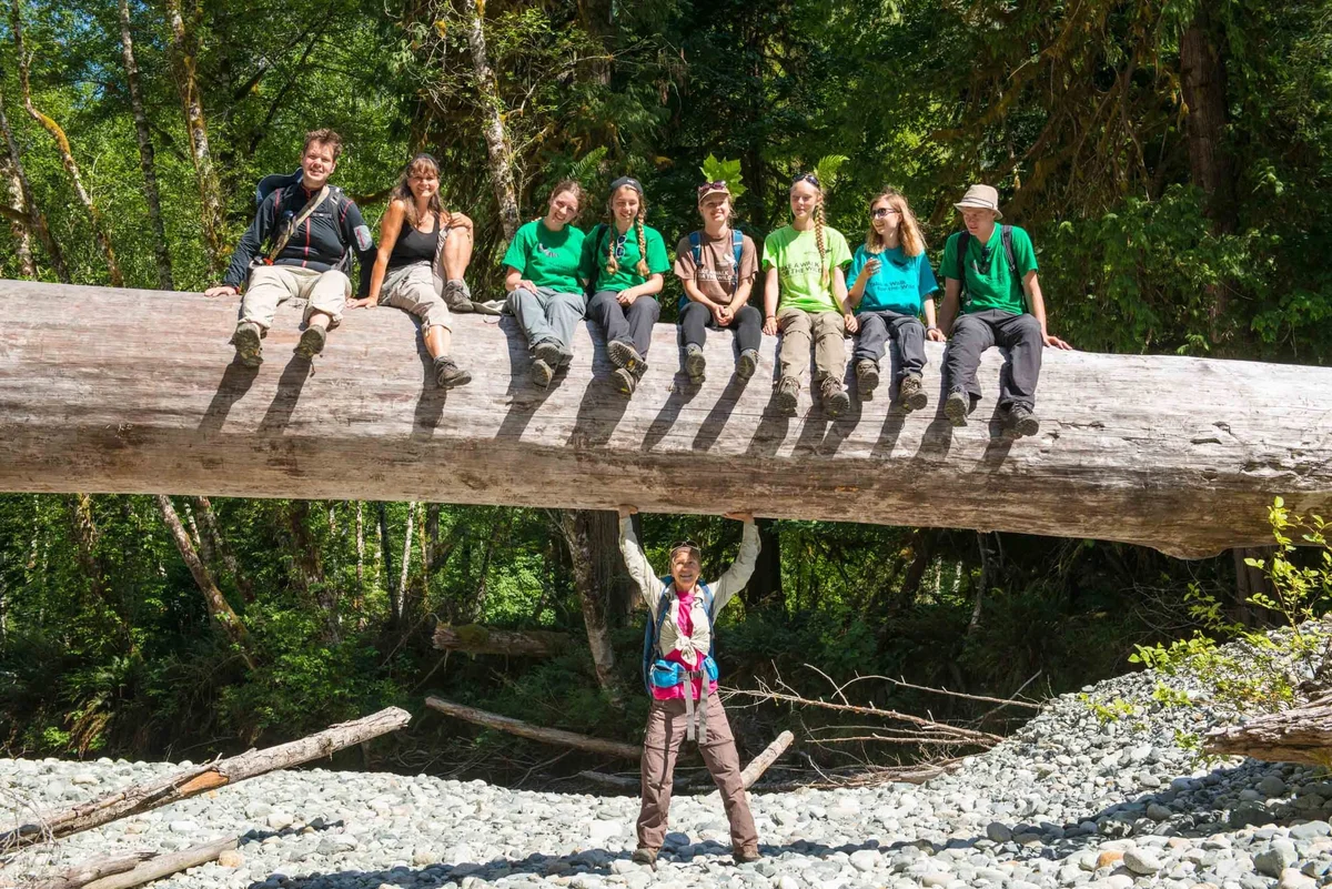 Group of youth sitting on a fallen log in the forest, with one person standing underneath with arms raised.