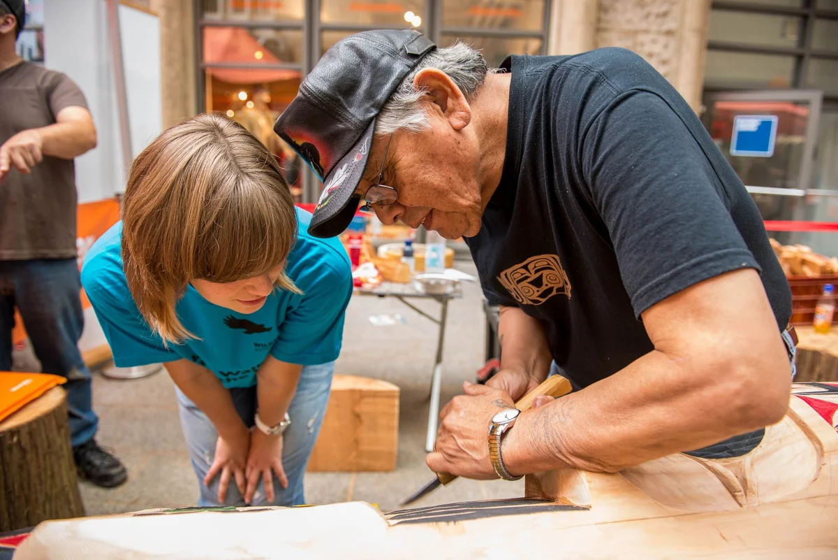 An older man in a cap demonstrates woodworking techniques to a young child at a workshop table.