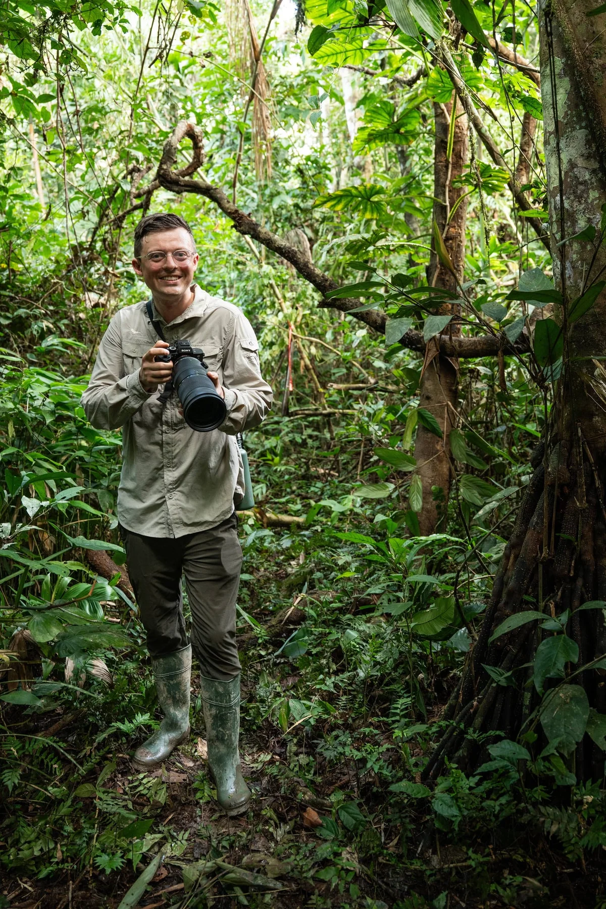 Smiling man holding a telephoto lens camera in a dense rainforest, wearing khaki shirt and boots.