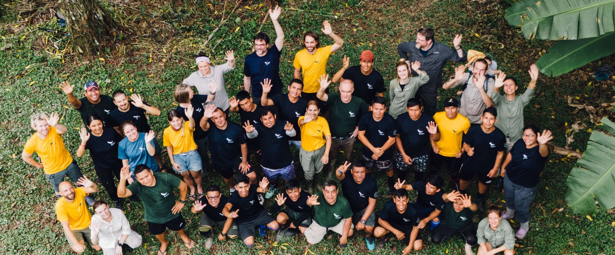 Aerial view of a large group of people in yellow and black shirts posing cheerfully together on green grass.