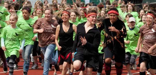 Large group of children and adults running together on a red track during a community athletic event.