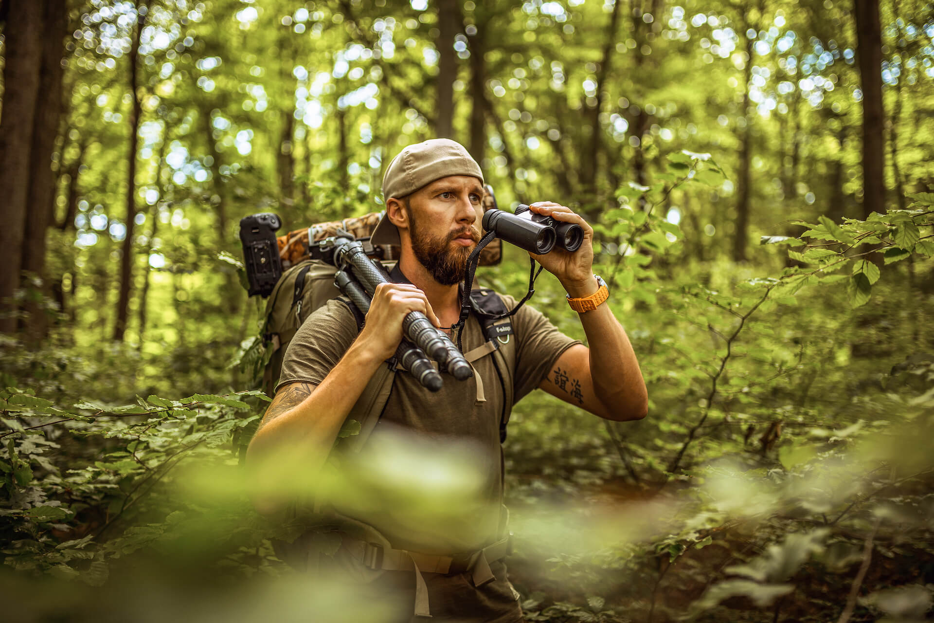 Bearded man wearing cap looks through binoculars in dense forest with backpack and camera gear.