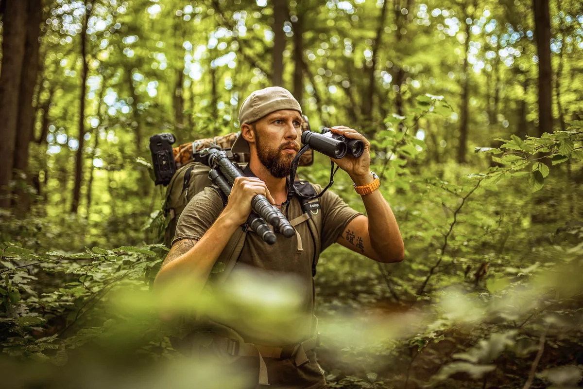 Bearded man wearing cap looks through binoculars in dense forest with backpack and camera gear.