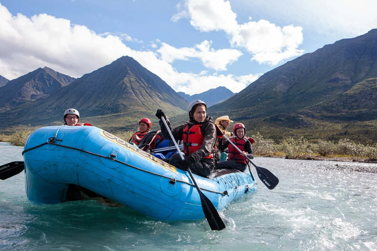 Group of five people wearing helmets in a blue inflatable raft on a turquoise river, surrounded by mountains.