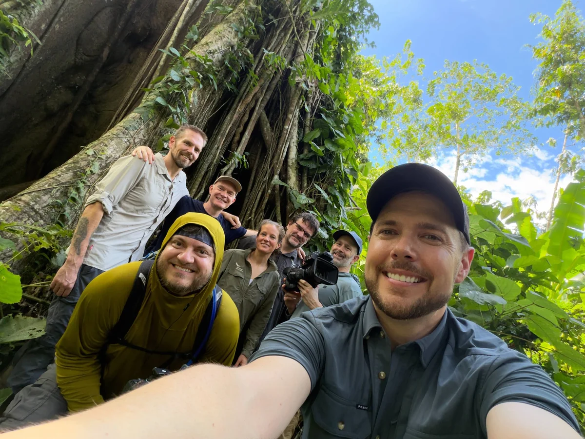 Group of smiling men posing together in front of a massive ancient tree with lush foliage in a tropical forest.