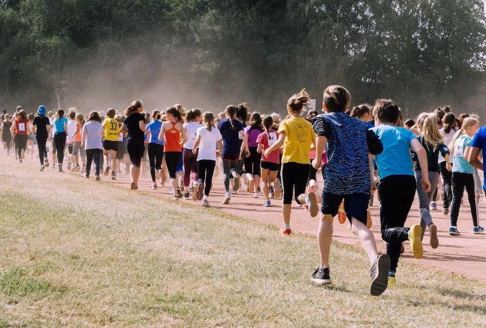 Large group of children and youth running on a red track surrounded by trees with dust clouds rising.