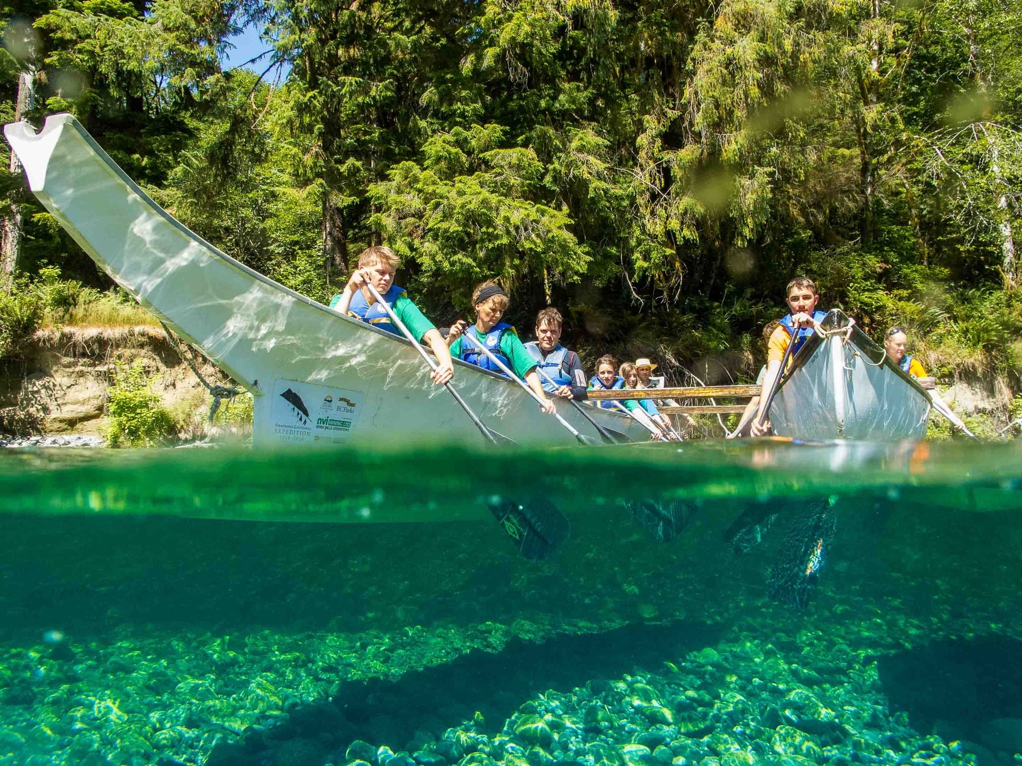 Children sitting in a glass-bottom boat over clear green water, viewing fish and aquatic plants beneath the surface.