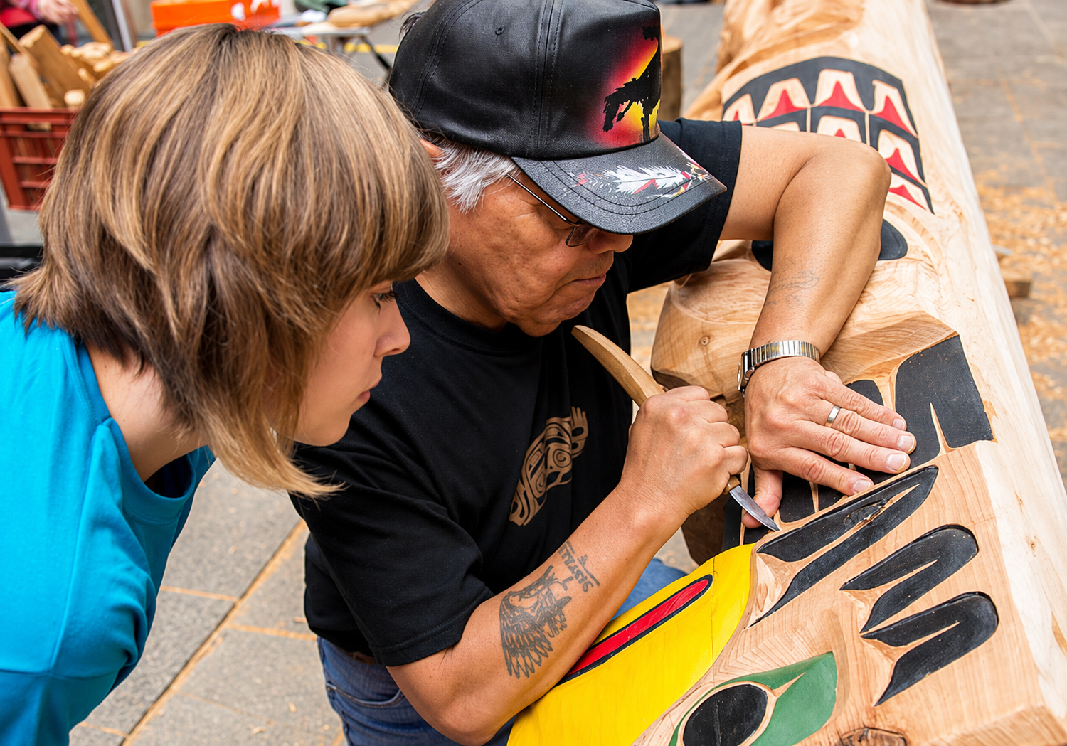 Woodcarver wearing a baseball cap demonstrates traditional carving techniques to a young observer while working on a wooden totem pole.