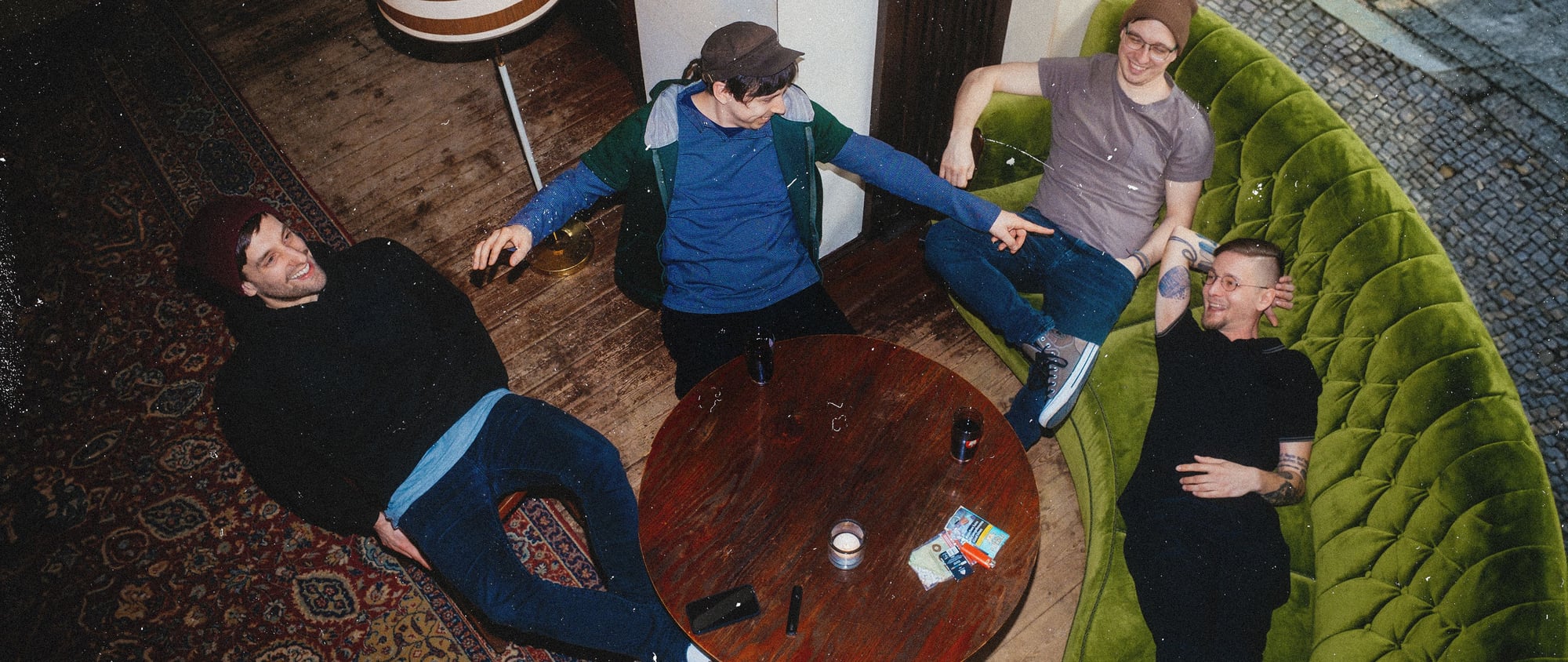 Cuatro hombres sentados alrededor de una mesa de madera en una sala con sofá verde.