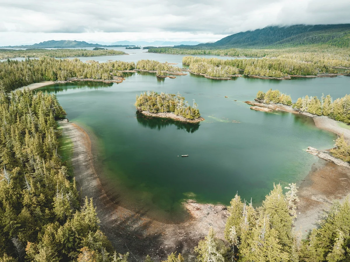 Aerial view of turquoise lake with forested islands, surrounded by dense coniferous forests and mountains under cloudy skies.