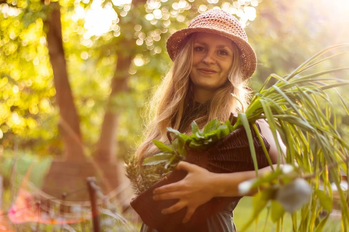 Girl wearing a pink hat holding a potted plant in a sunny garden with trees in the background.