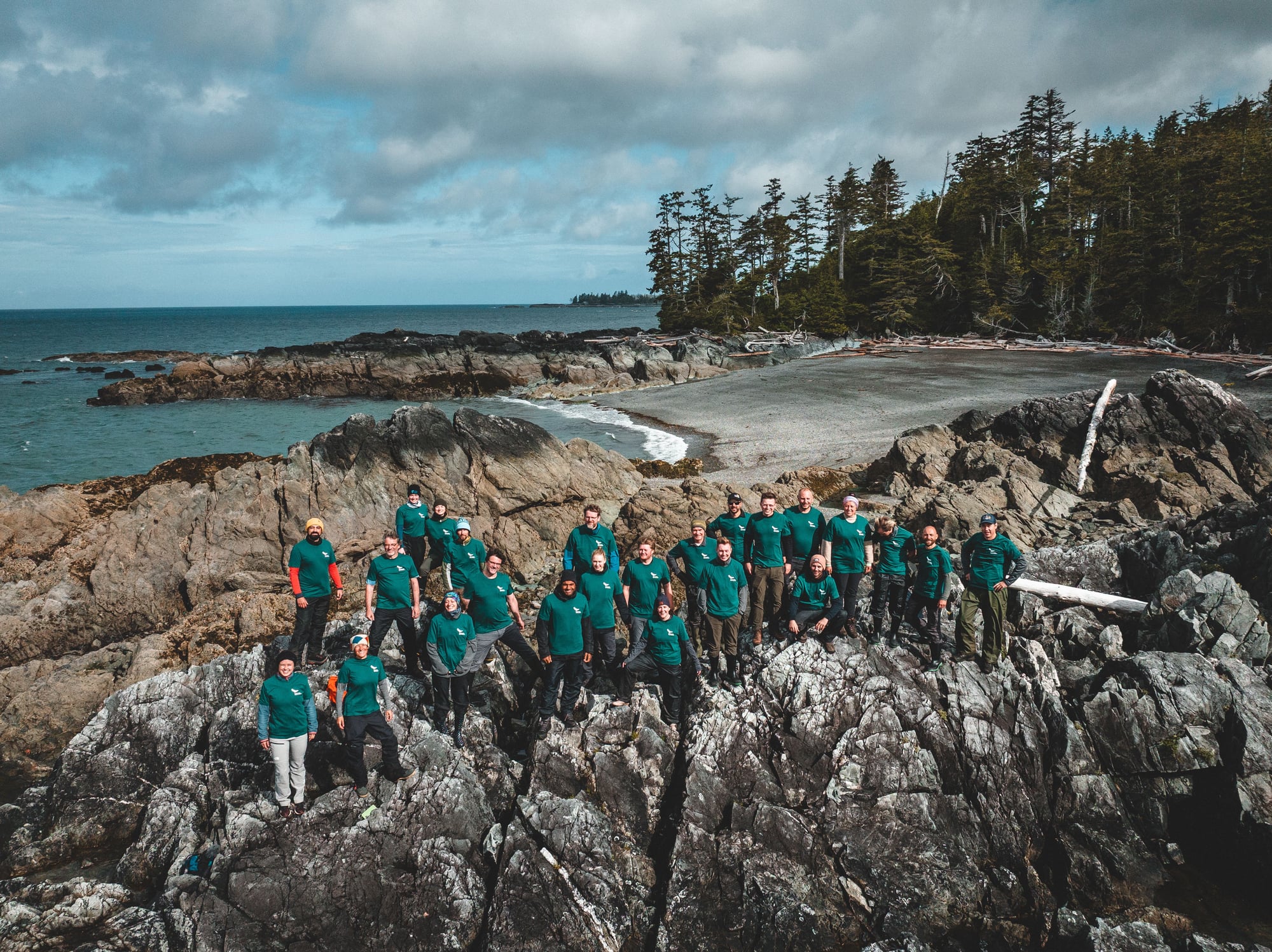 Large group of people wearing matching teal shirts posing on rocky shores beside a beach and evergreen forest.