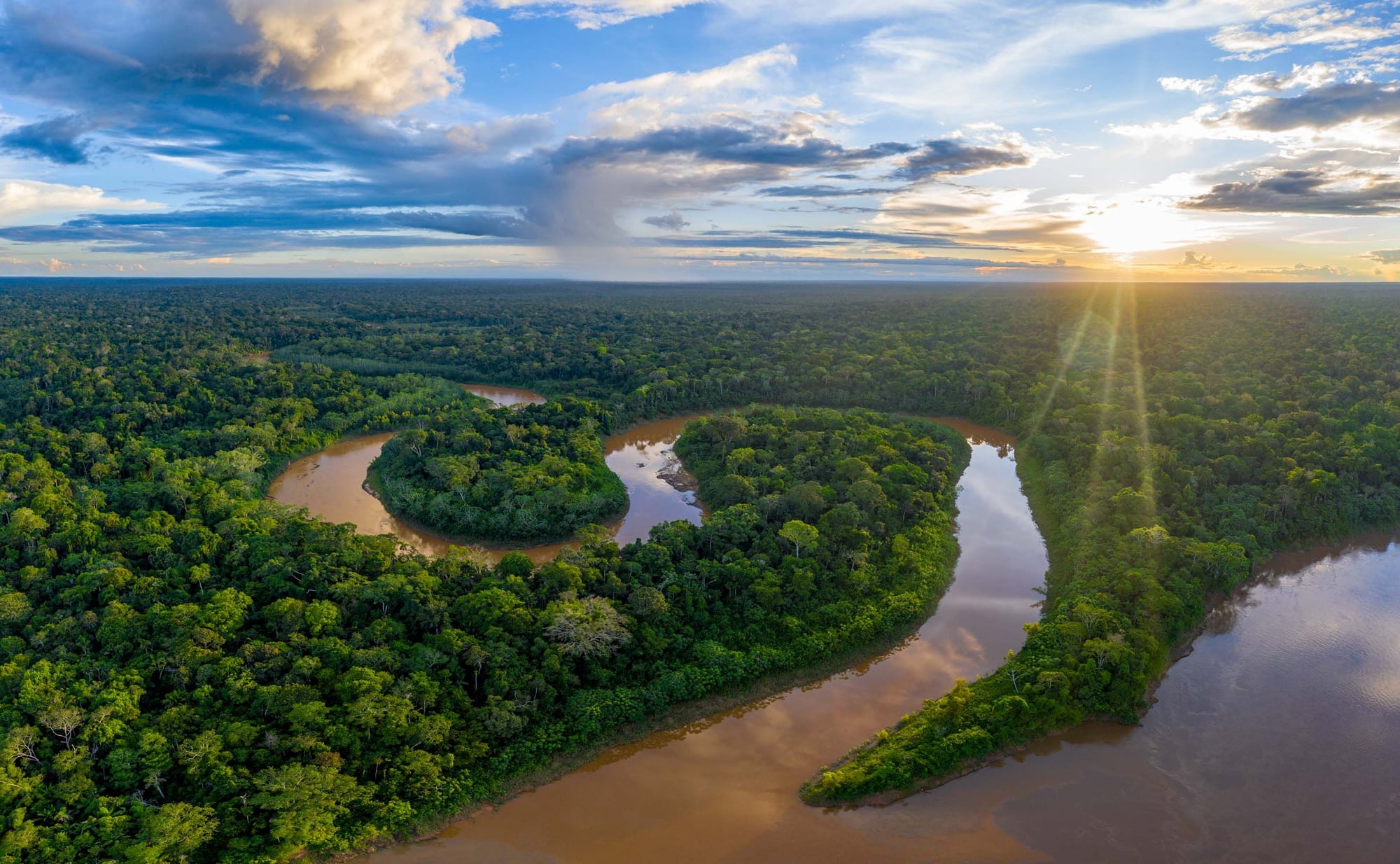 Aerial view of Amazon rainforest with winding rivers, lush green canopy, and dramatic sky at sunset with golden light rays.