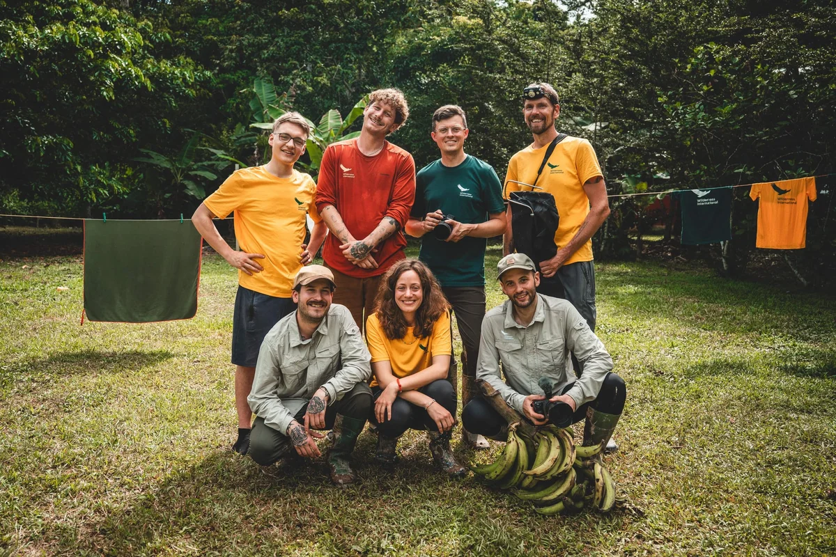 Nueve personas sonrientes con ropa de outdoor posan en un jardín con plátanos y ropa tendida.