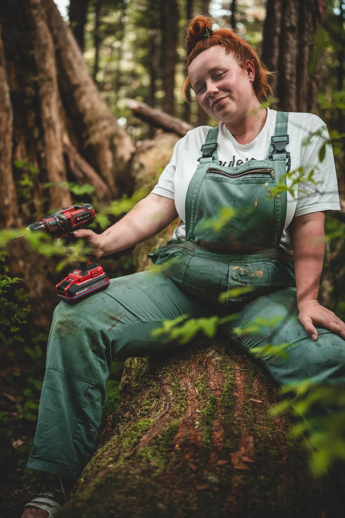 Red-haired woman in green work overalls holding a red cordless drill in a forest setting.