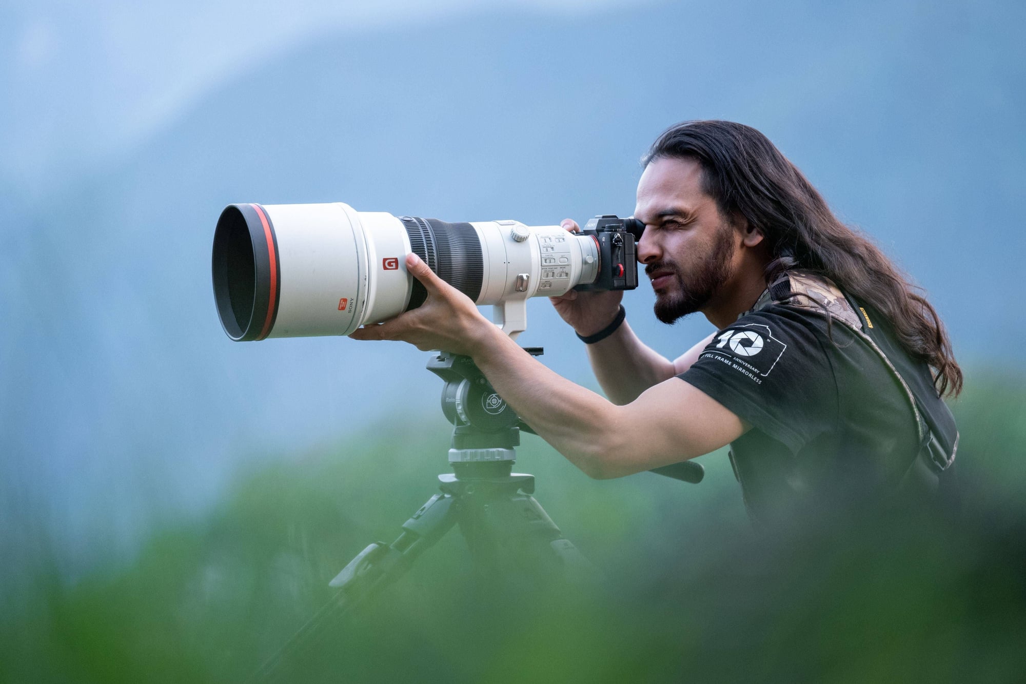 Fotógrafo con cabello largo oscuro usando un teleobjetivo grande montado en trípode bajo cielo azul.