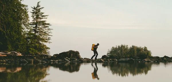 Un senderista con mochila amarilla salta entre rocas sobre un lago tranquilo, rodeado de bosque.