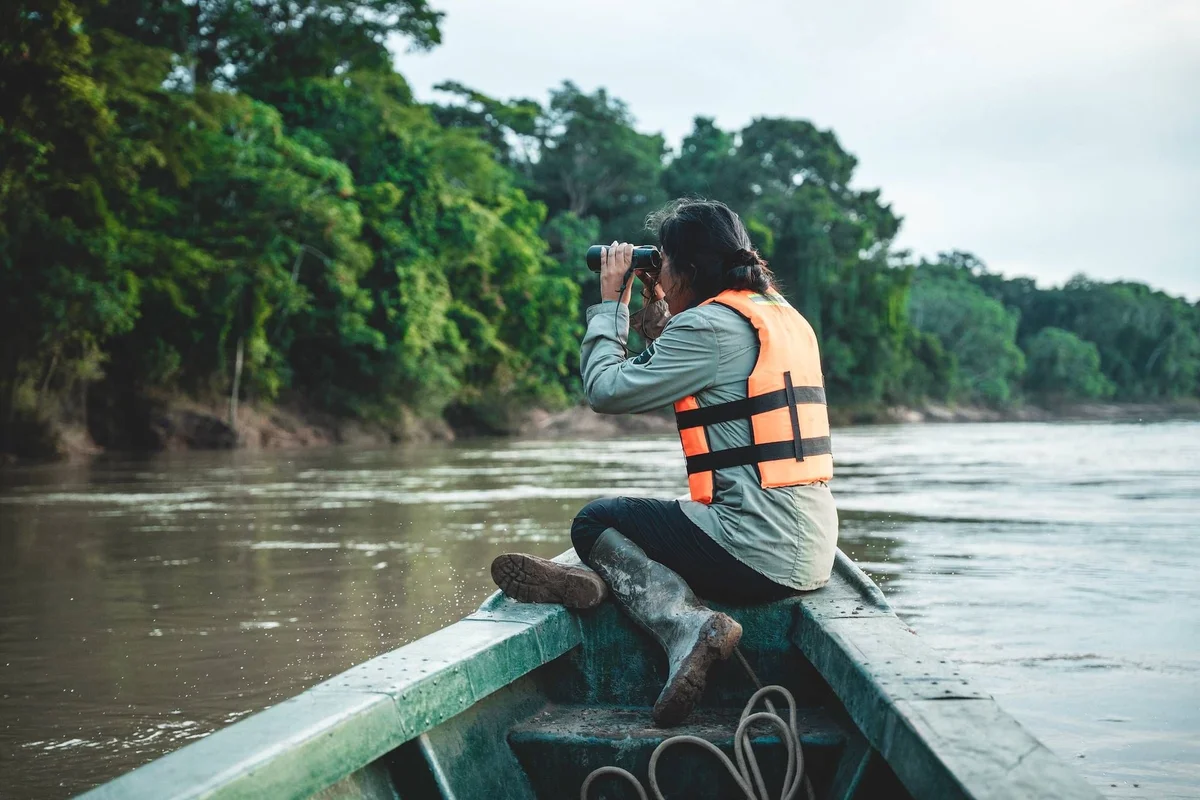 Person wearing orange life vest sits in boat on river, looking through binoculars at lush green forest