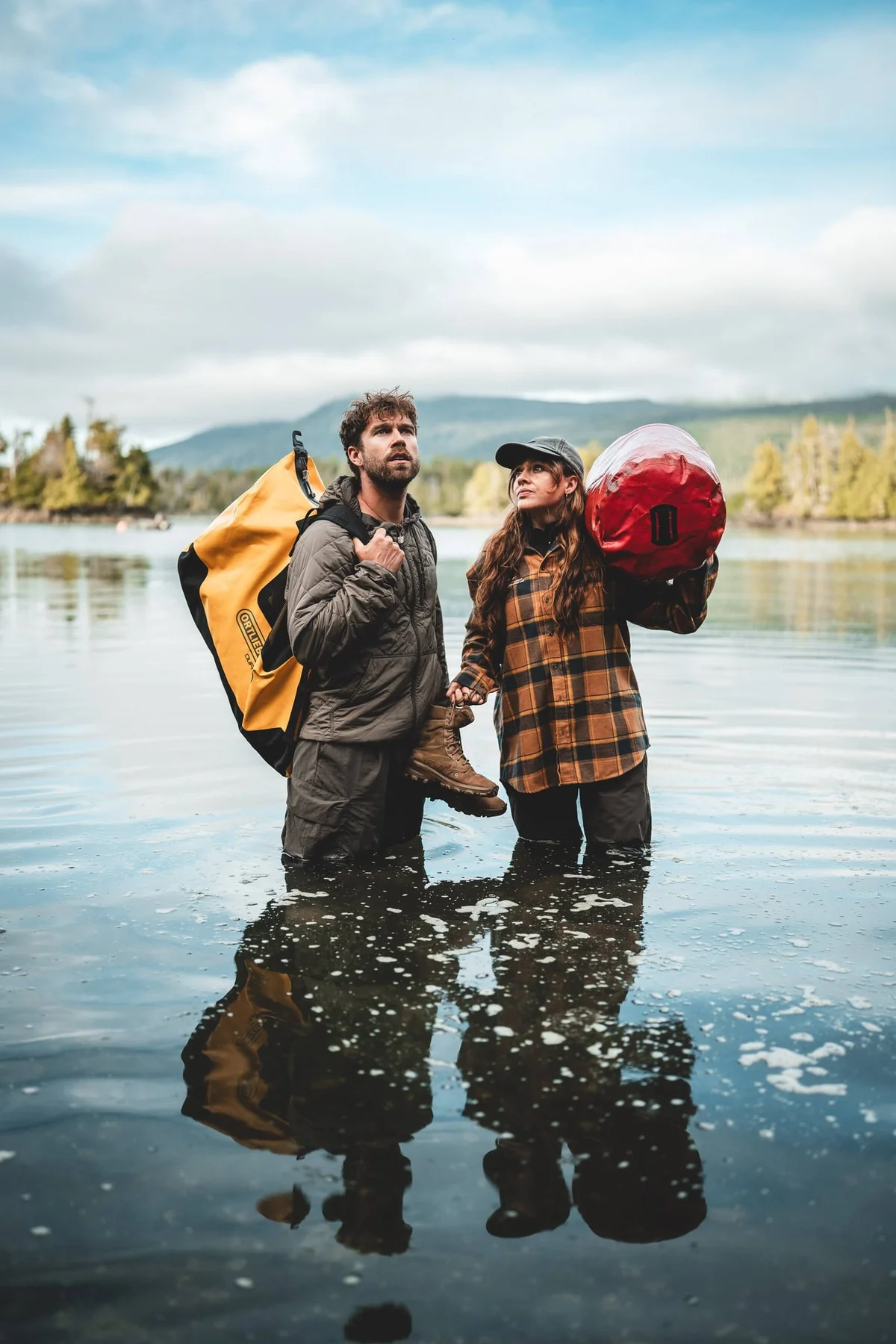 Two hikers stand in shallow water of a mountain lake with snowy peaks visible in the distance behind them.