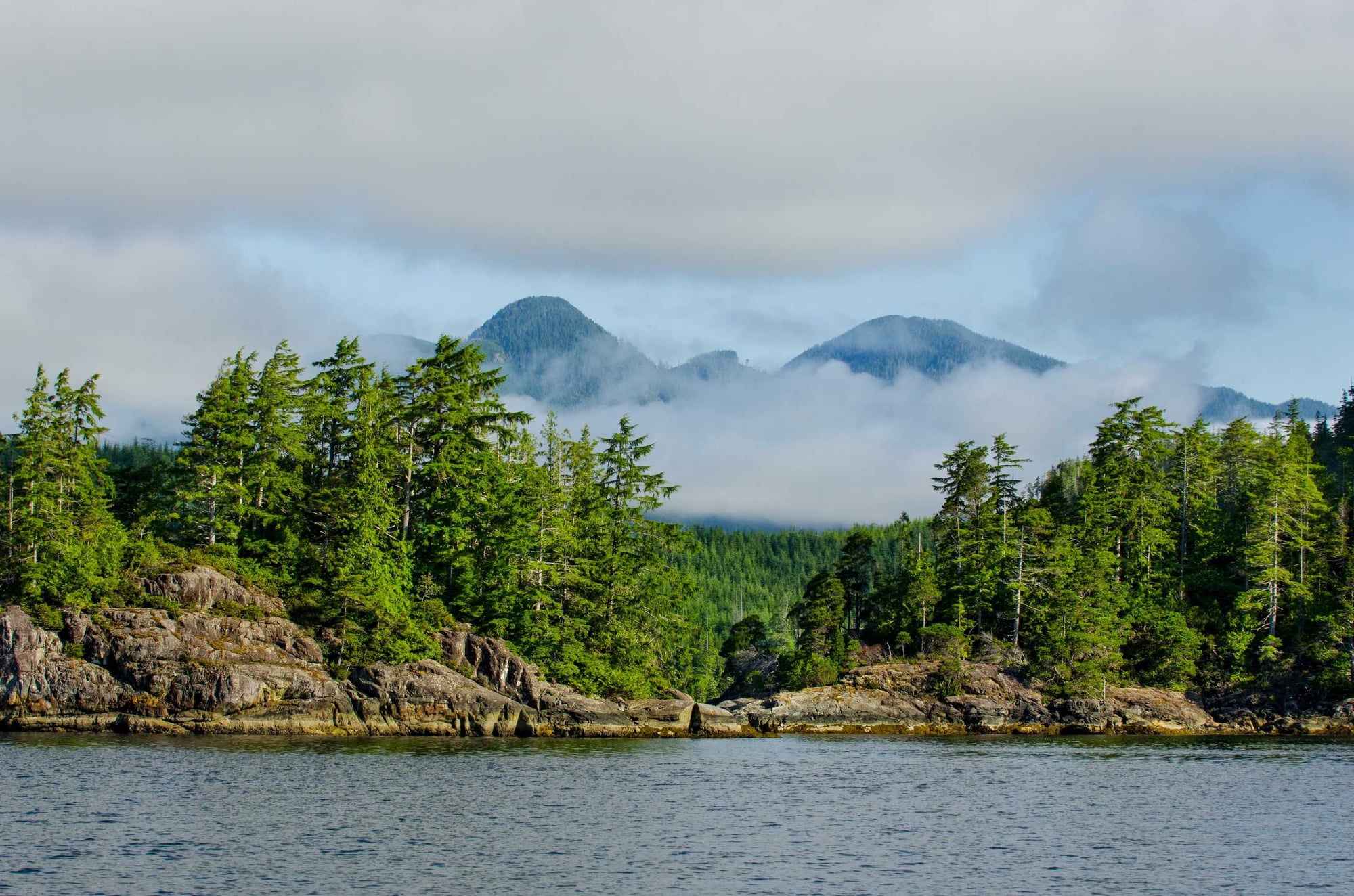 Mountain landscape with two snow-capped peaks, evergreen forest shoreline, and rocky outcrops along a lake.