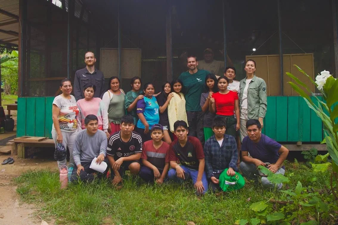 Group of approximately 20 people of varying ages posing together in front of a green wooden building with a garden.