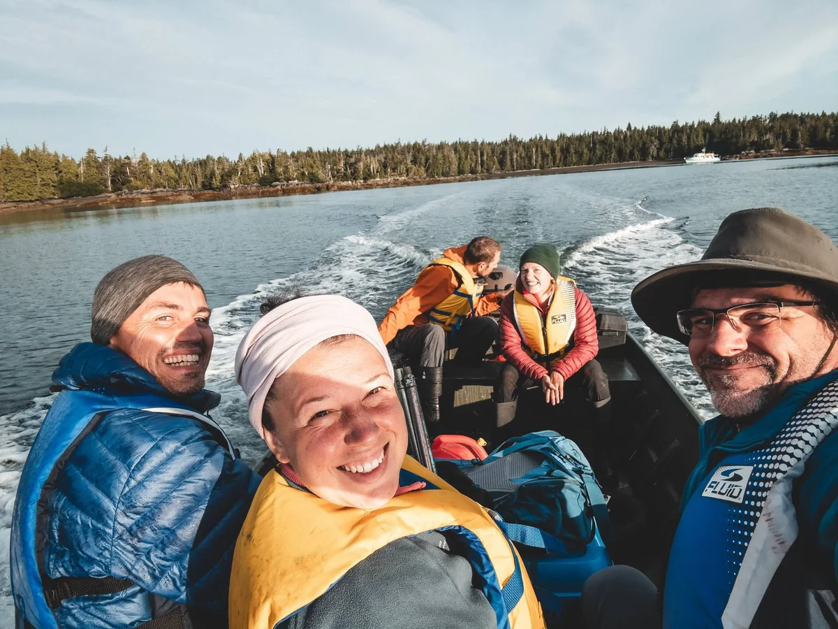 Group of five people wearing life jackets on a speedboat on a lake, with forested shoreline and mountains in the background.