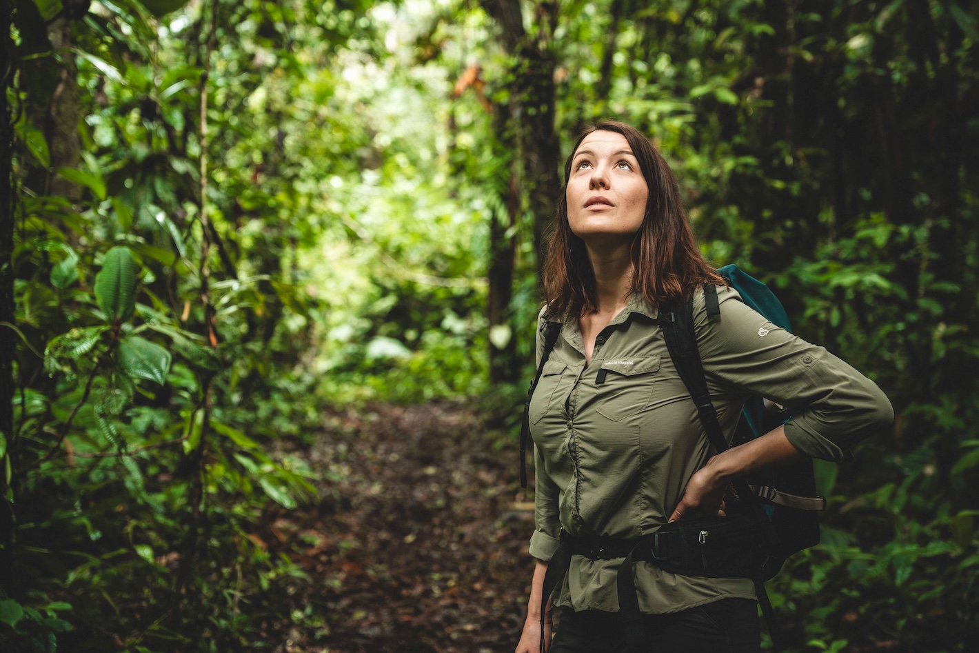 Mujer con mochila mira hacia arriba entre el follaje verde denso del bosque.