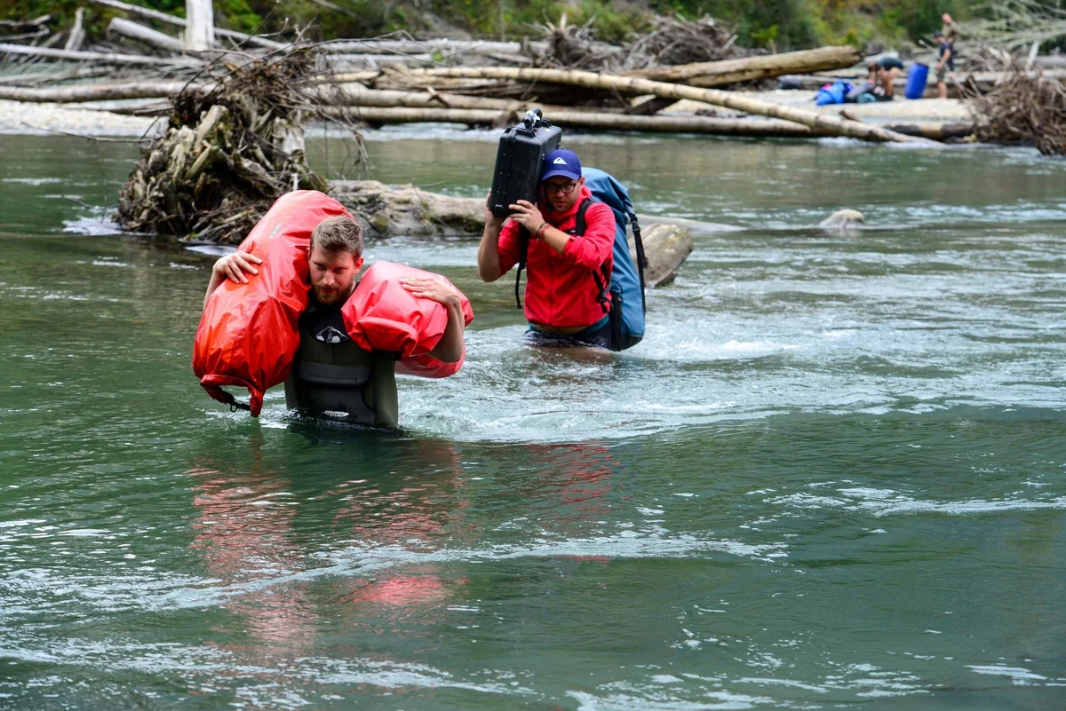 Dos personas con chalecos salvavidas rojos vadean un río cargando equipaje, con troncos flotantes al fondo.