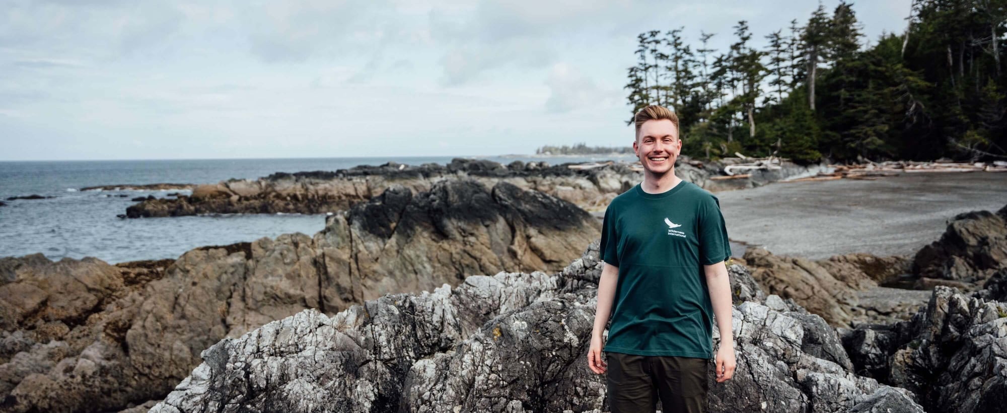 Young man in dark green shirt standing on rocky coastal rocks with ocean and forest backdrop