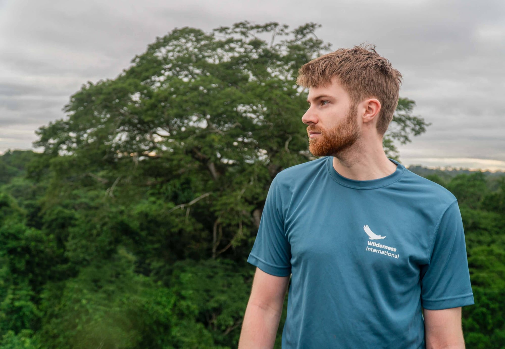 Bearded man in blue Wilderness International shirt gazes left against lush green forest backdrop.