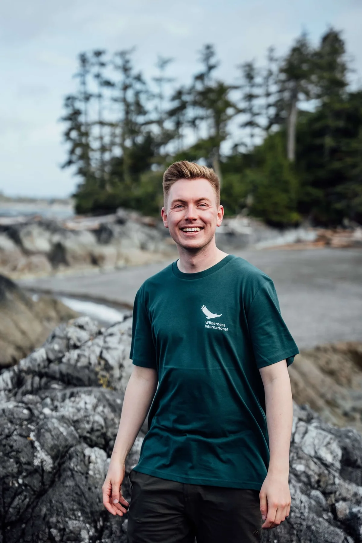 Smiling man in teal Wilderness International shirt stands on rocky coastal terrain with forested hills and water in background.