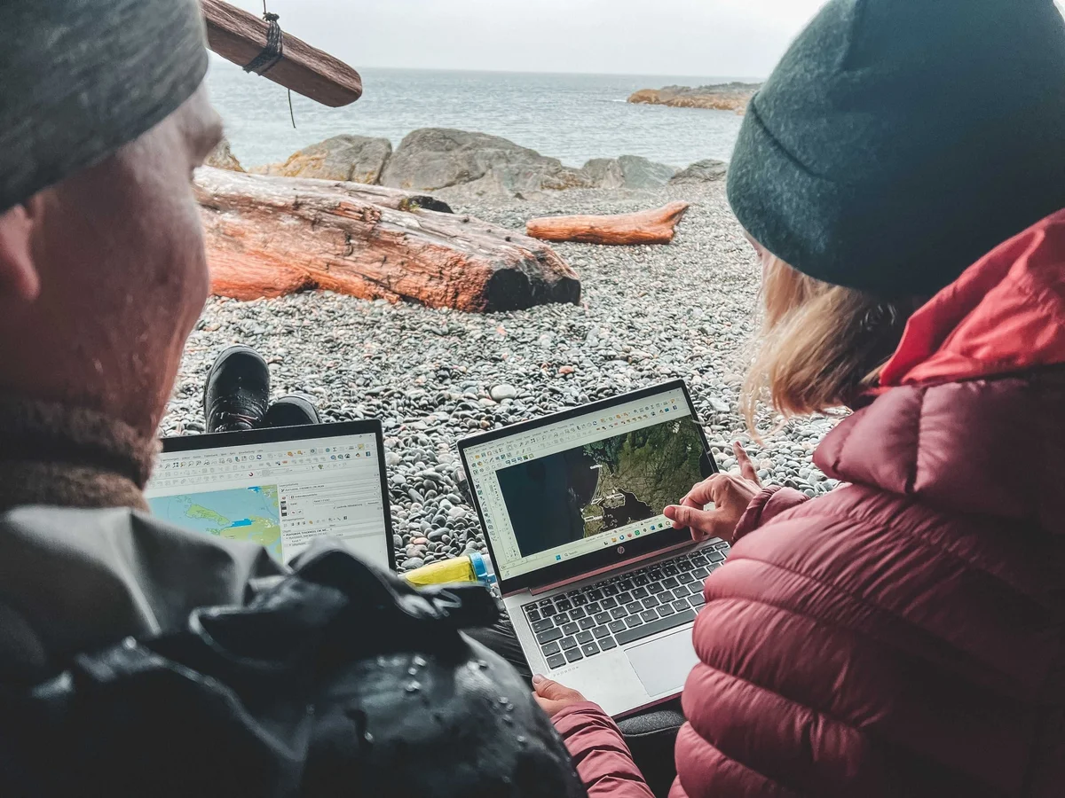 Two people working on laptops on a rocky beach with driftwood and ocean view.