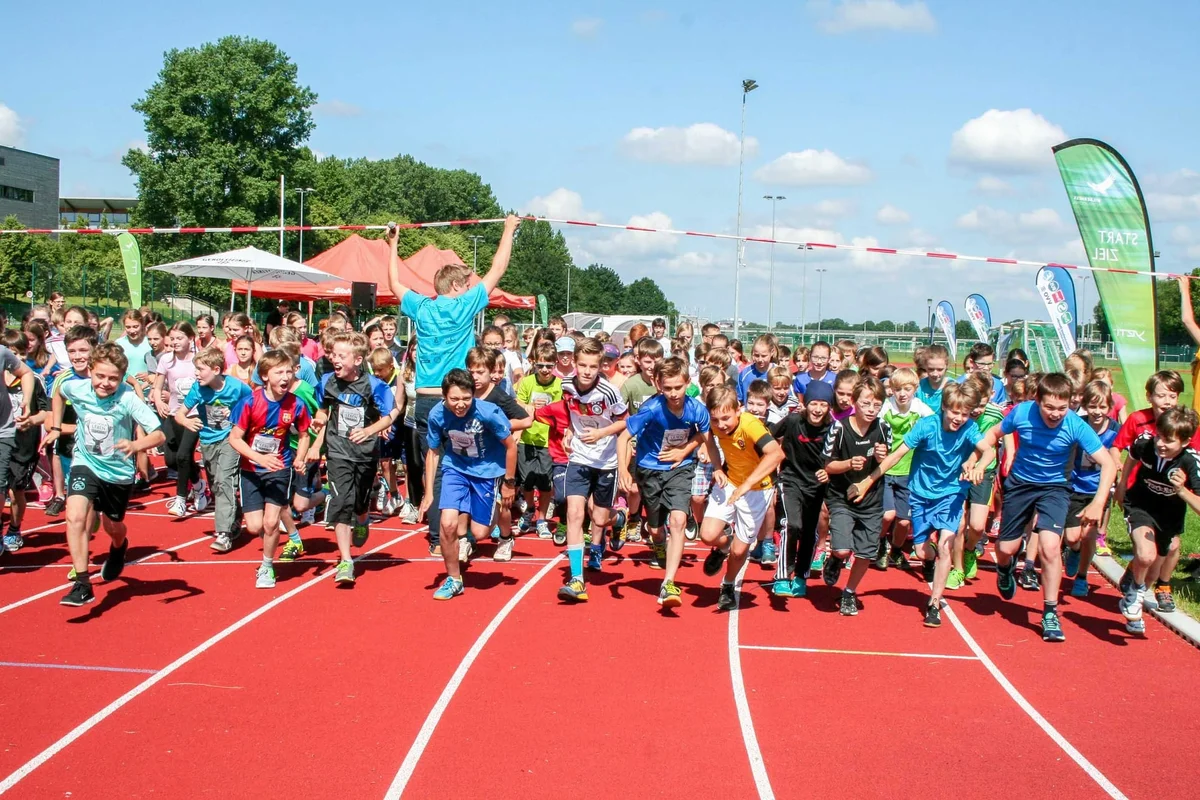 Large group of children running on a red athletic track during an outdoor children's sports event on a sunny day.