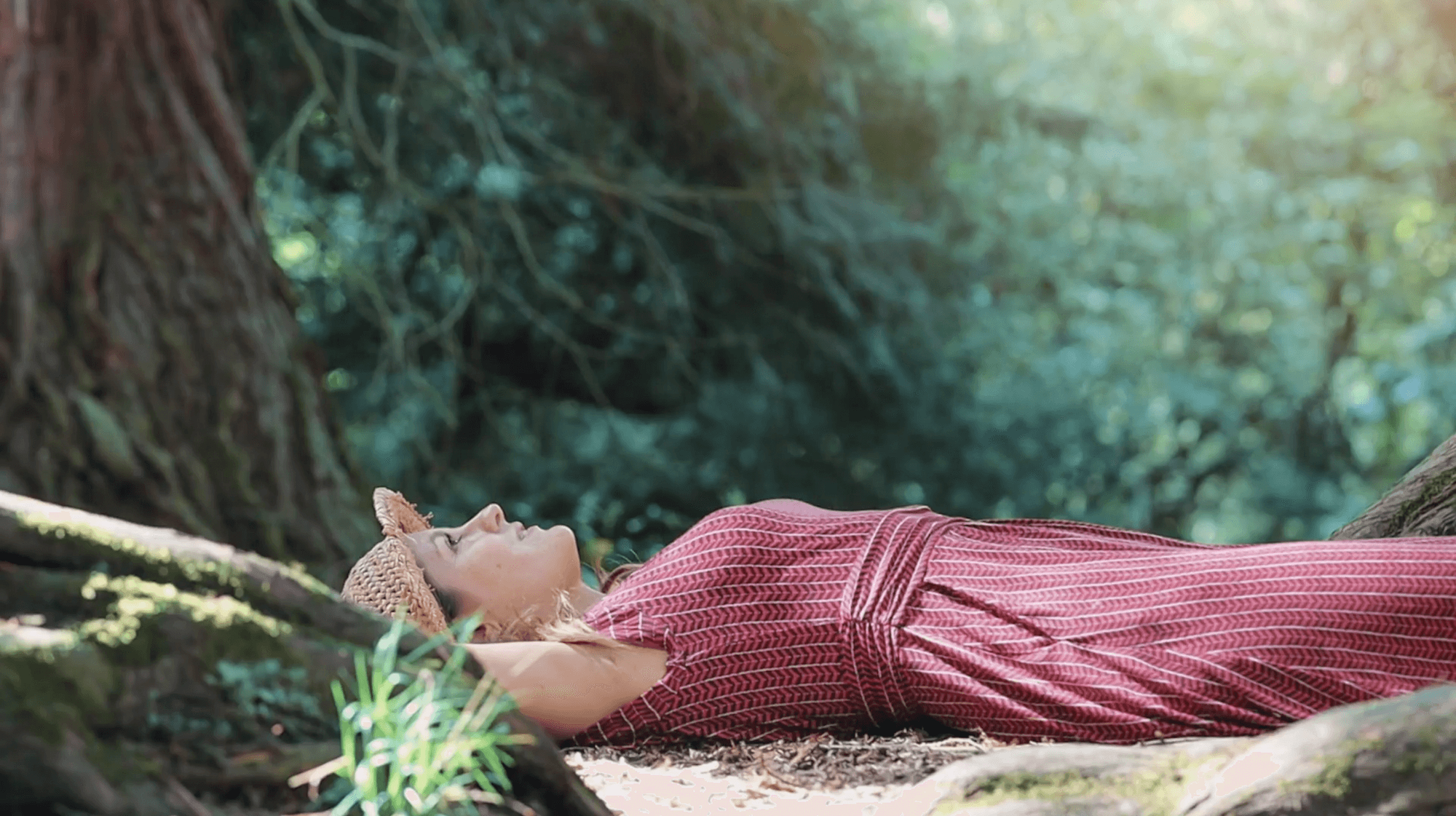 Woman in pink knit dress resting on forest floor surrounded by moss-covered logs and greenery.