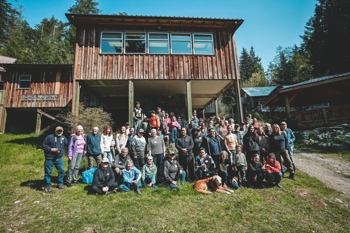 Large group of people posed in front of a modern wooden structure on a grassy field with coniferous trees in the background.