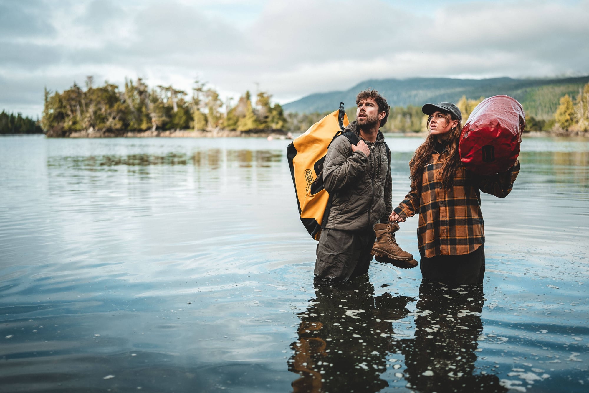 Two hikers with backpacks wade through a lake surrounded by forested mountains under a cloudy sky.