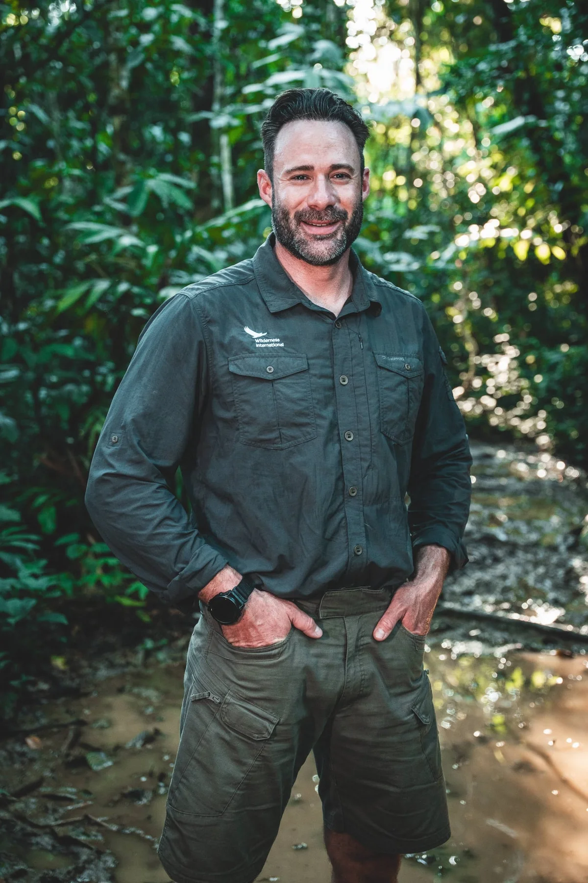 Hombre sonriente con uniforme de trabajo oscuro de pie en un entorno selvático exuberante con vegetación verde.