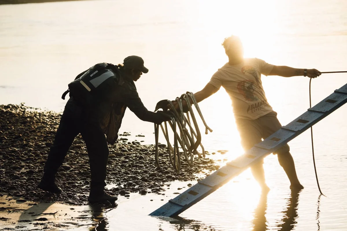 Dos buzos descargando equipos de buceo en la playa al atardecer.