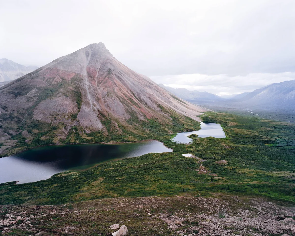 Volcanic cone with lava flows beside an alpine lake in a verdant valley under cloudy skies.