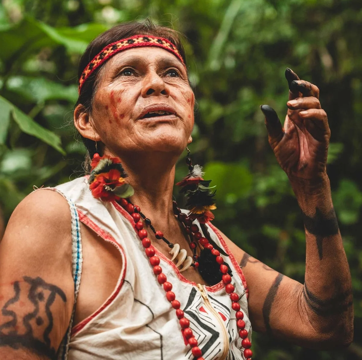 Indigenous man with facial paint, red beads, traditional jewelry, and headband gesturing in forest.