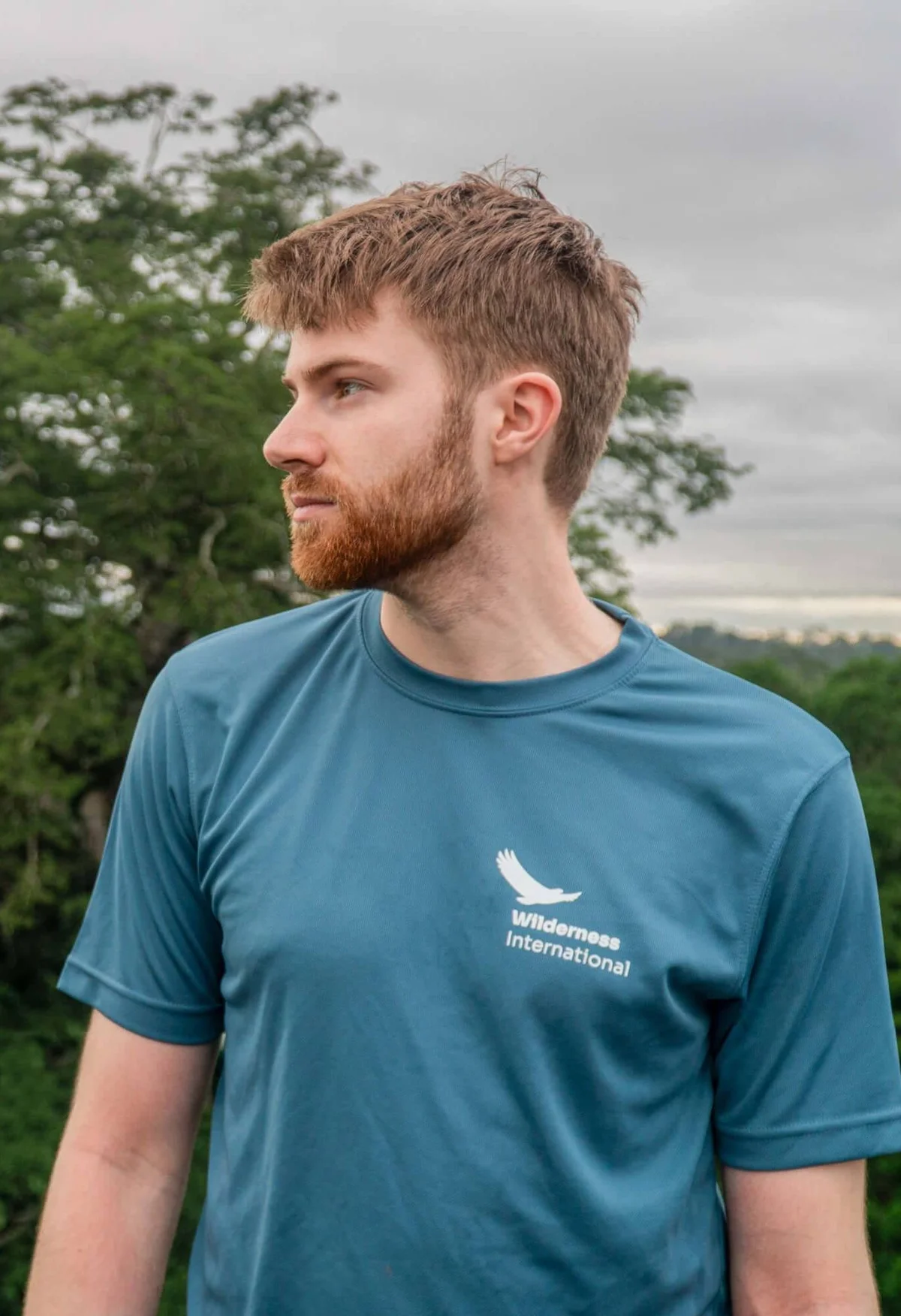 Bearded man in blue Wilderness International shirt looking to the side with green landscape and trees behind