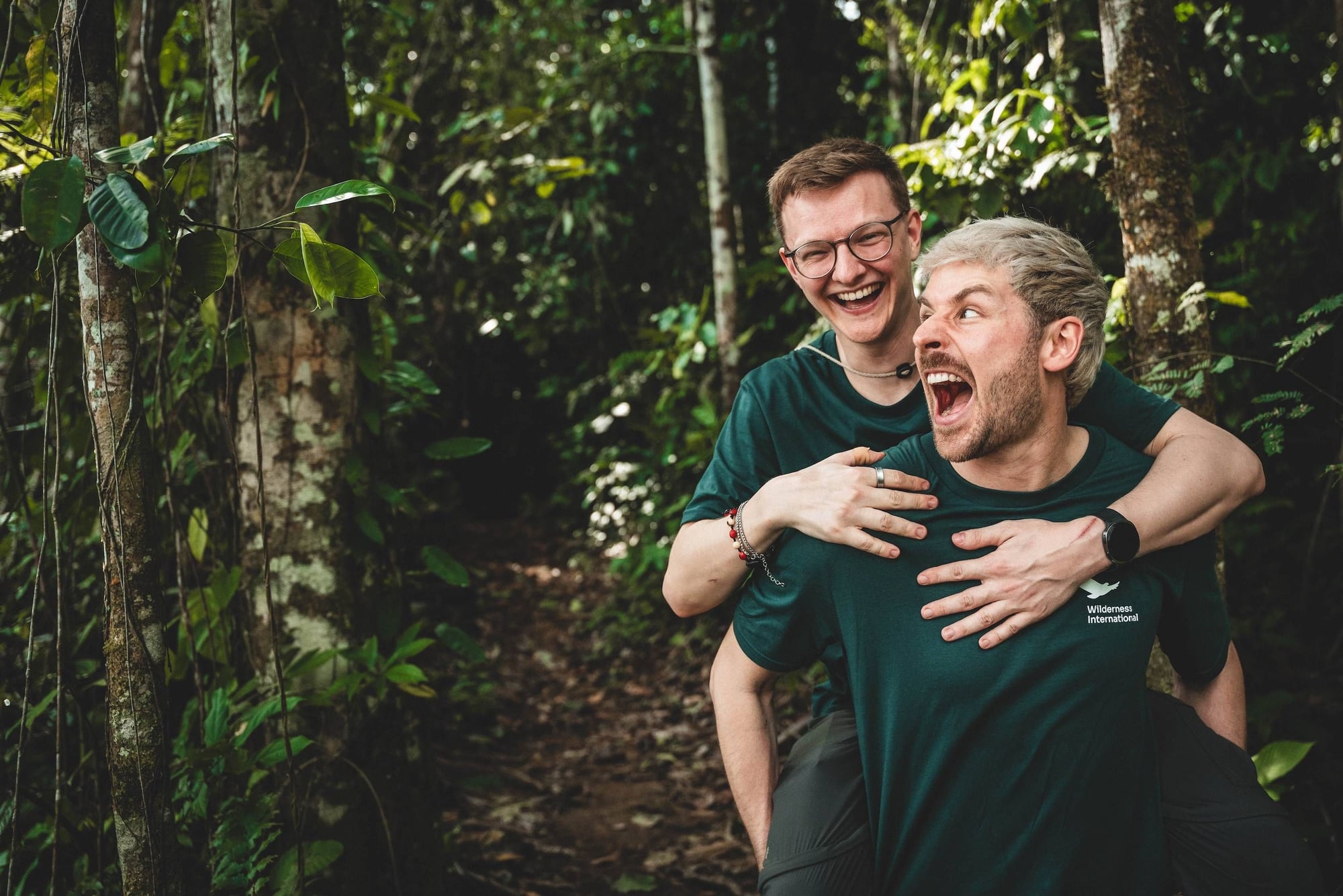 Dos hombres sonrientes en la selva, uno cargando al otro en la espalda. Ambos visten camisetas verdes de Wilderness International.