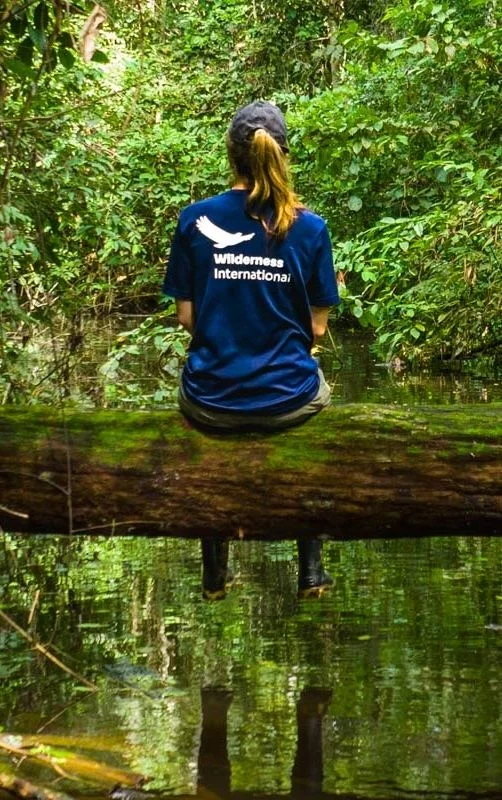 Person sitting on log over green water wearing blue Wilderness International t-shirt