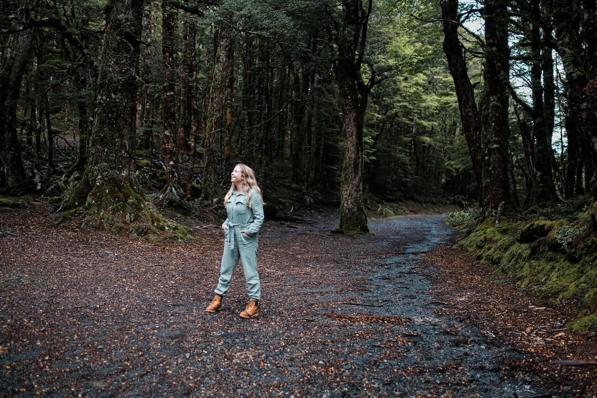 Woman in light blue suit standing on forest trail between moss-covered trees, looking upward