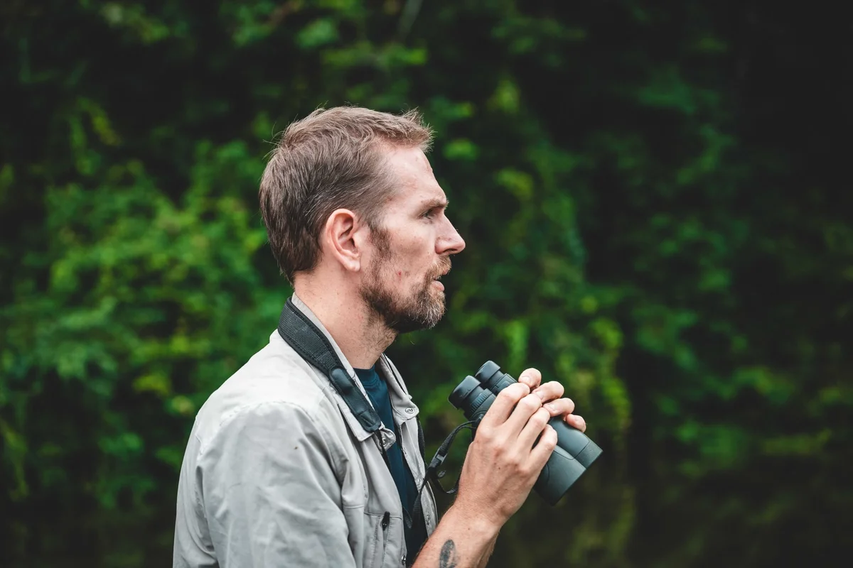 Bearded man in profile holding binoculars against lush green foliage background.