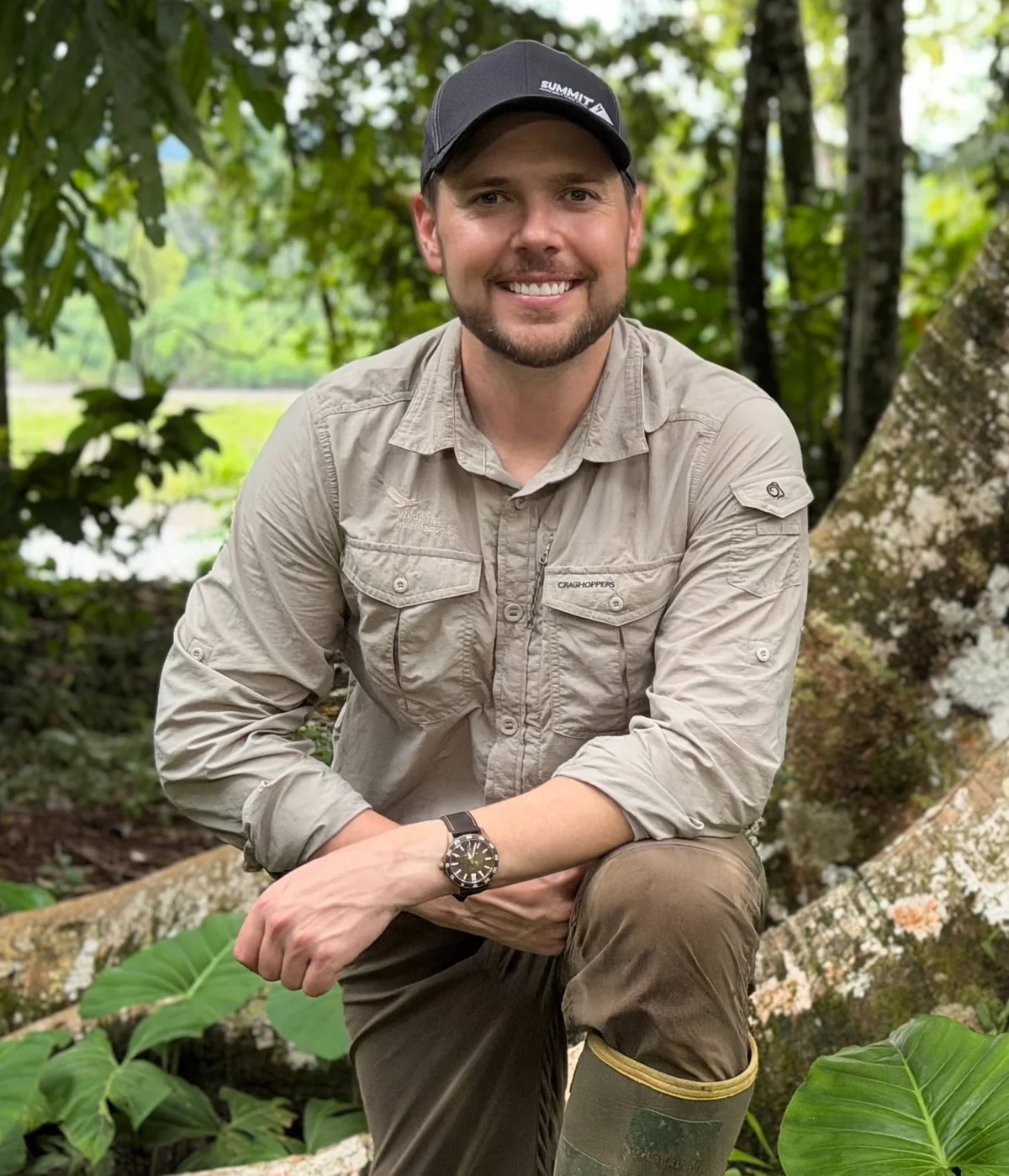 Smiling man in khaki outdoor gear and baseball cap crouching on a mossy log surrounded by green forest foliage