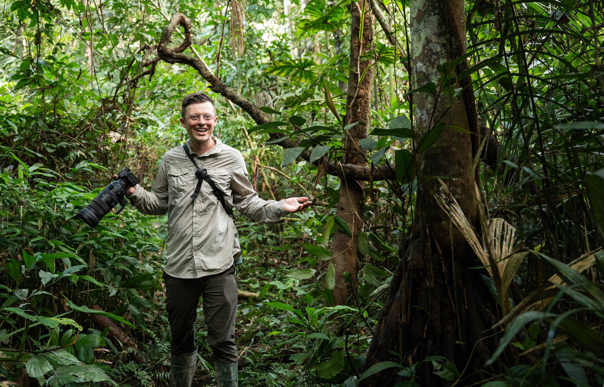 Smiling man holding camera in dense rainforest surrounded by lush green vegetation and large trees.