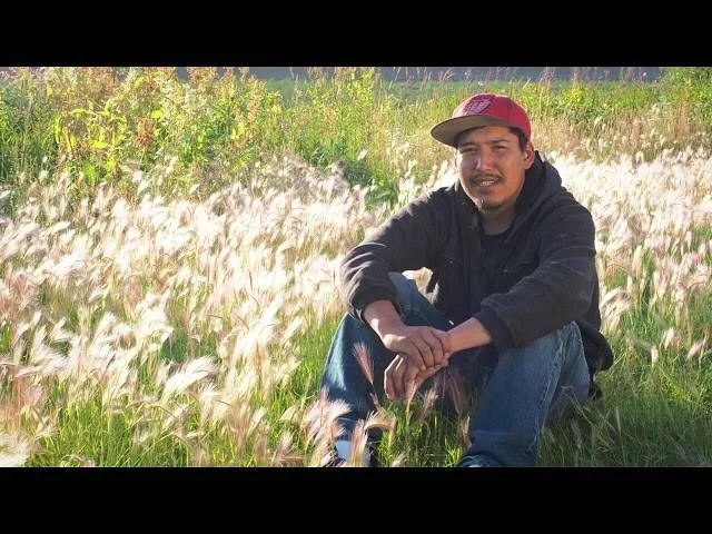 Man wearing a red cap and dark jacket crouching in a field of white wildflowers.