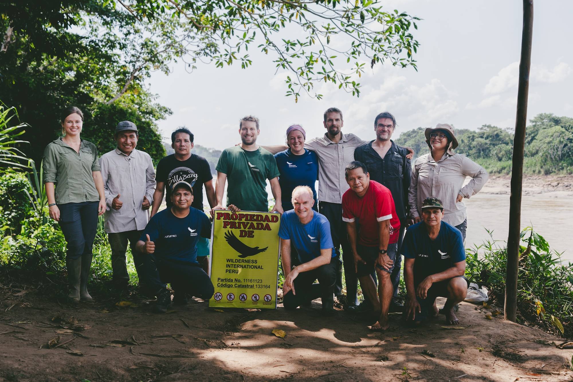 Group of people holding Wilderness International sign at riverbank in Peru. Diverse team poses together outdoors.