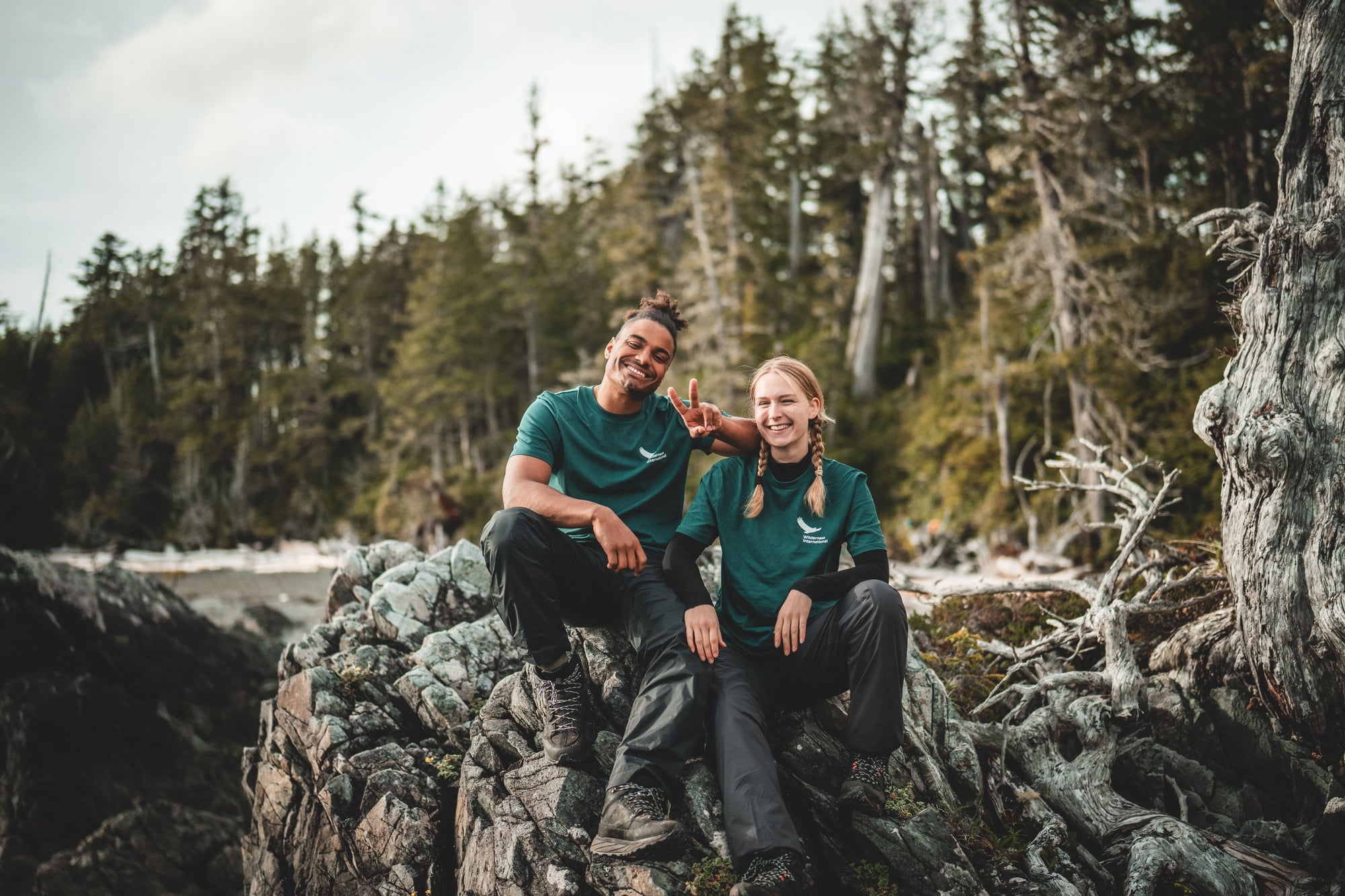 Dos personas sonrientes con camisetas verdes sentadas en rocas junto a un lago rodeado de bosque de coníferas